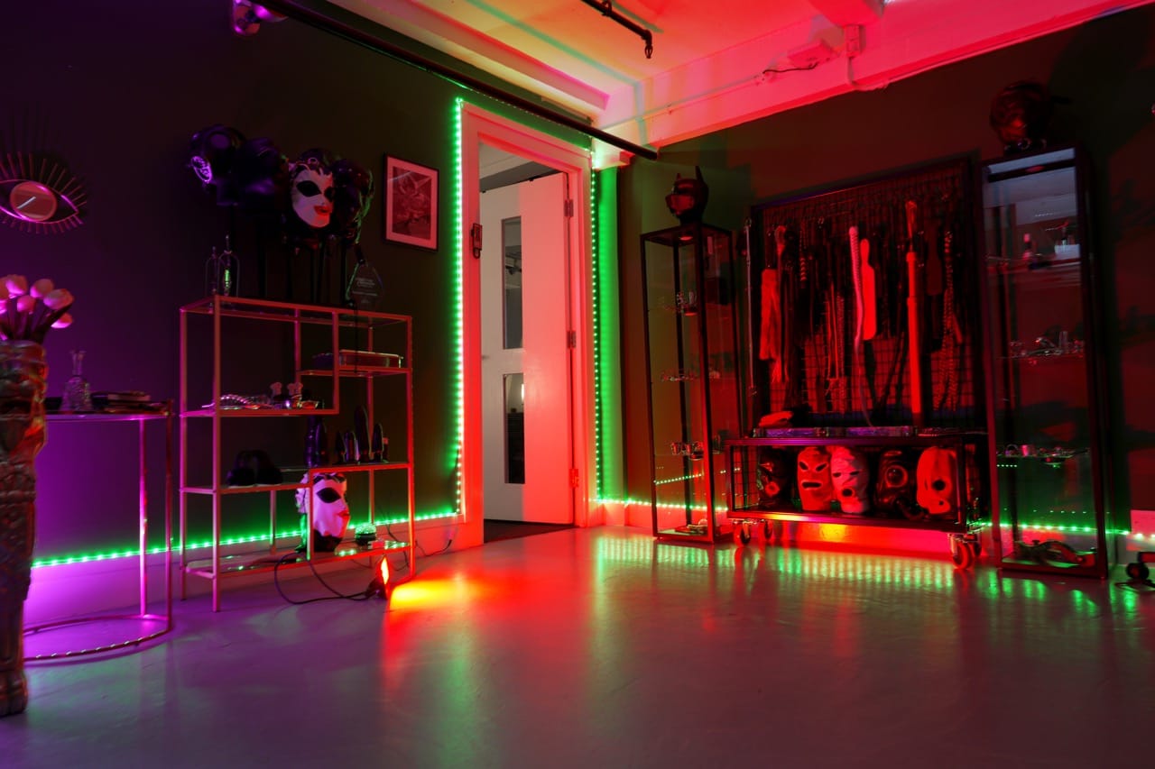 Wide view of the main chamber at London Dungeon Suite with display shelves, masks and a wall rack of floggers under green and red light
