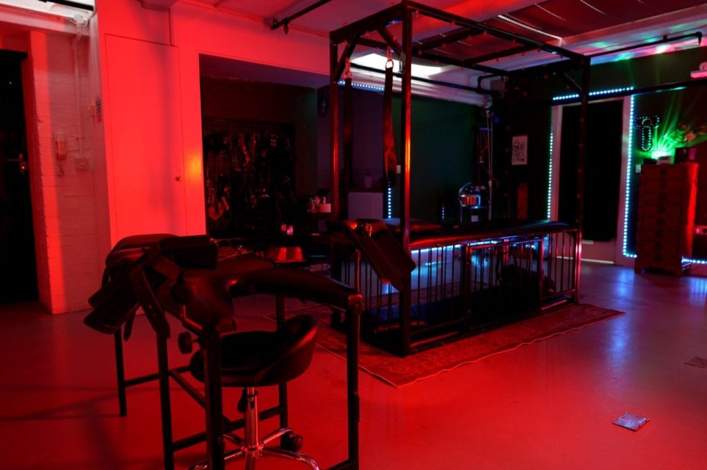 Gyno chair with leg supports in the foreground and the cage and bondage frame behind, under red light