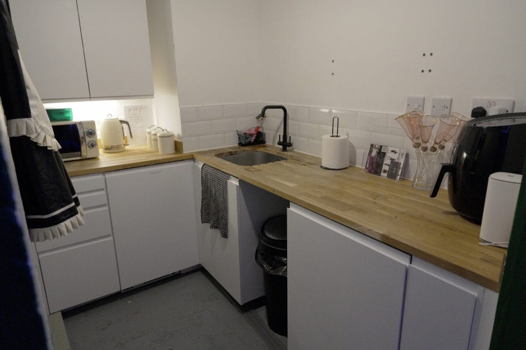 Compact kitchenette with white cabinets, a wooden worktop, sink, kettle, microwave and air fryer