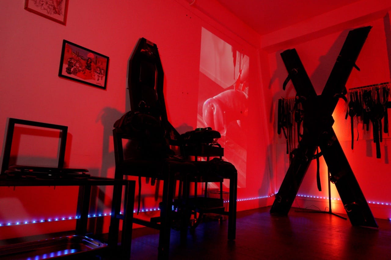 Freestanding St Andrew’s cross and dungeon furniture under red light at London Dungeon Suite